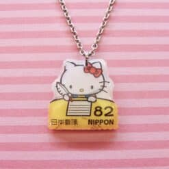 Hello Kitty postzegelketting met zilveren ketting en gele postzegelhanger met rood strikje en Japanse tekst.
