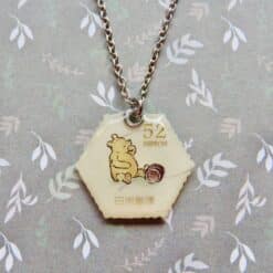 Crème kleurige zeshoekige postzegelketting met Winnie de Poeh en vintage stijl opdruk, aan metalen ketting.